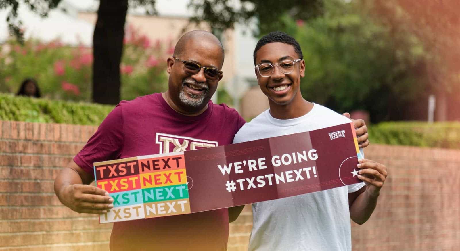 A parent and a student holding “WE’RE GOING #TXSTNEXT” signs on the Texas State campus